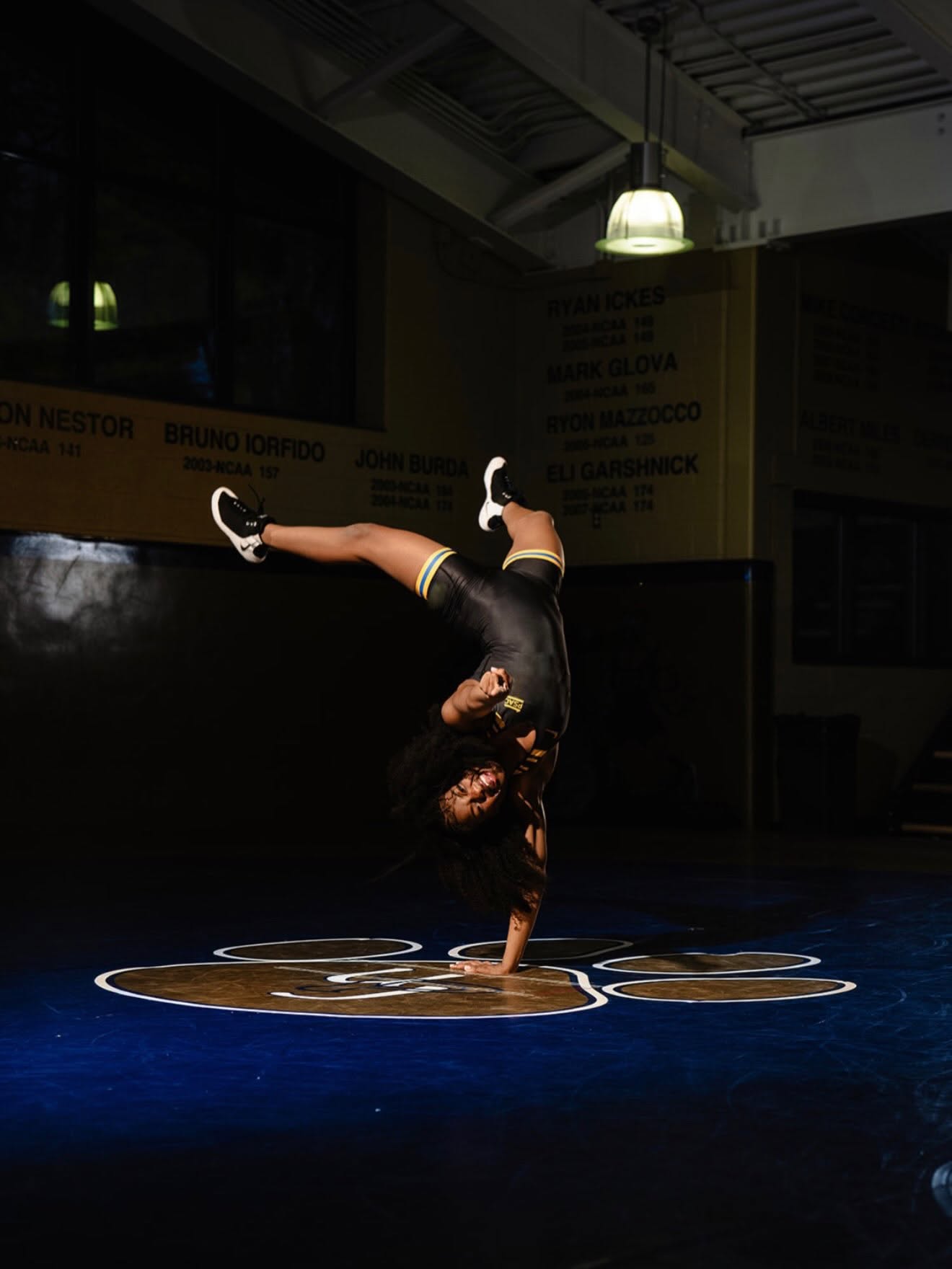 Tamara Humphries holding a one-handed handstand on the mat, lit by a single overhead lamp in an otherwise dark room.