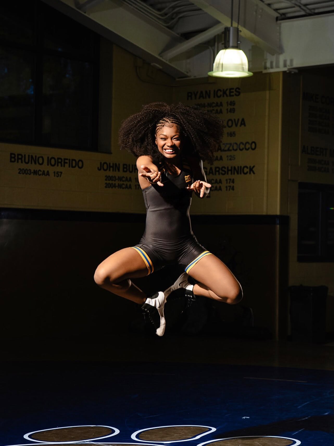 Tamara Humphries mid-air in a dark wrestling room, arms out and laughing, lit by a single overhead lamp, the wall of past qualifiers' names behind her.