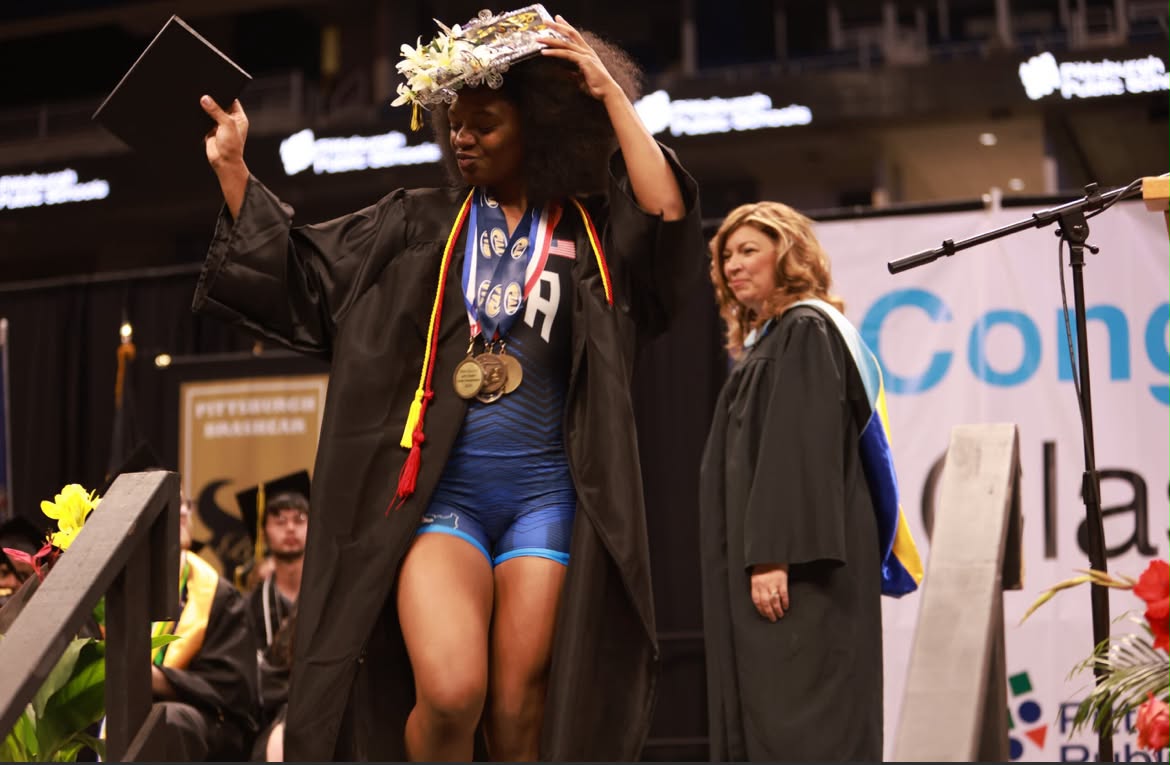 Tamara Humphries crossing the graduation stage at Brashear, wearing her Team USA singlet under an open gown with three medals around her neck.