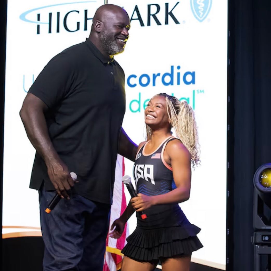 Tamara Humphries laughing on stage beside Shaquille O'Neal, wearing a Team USA singlet.